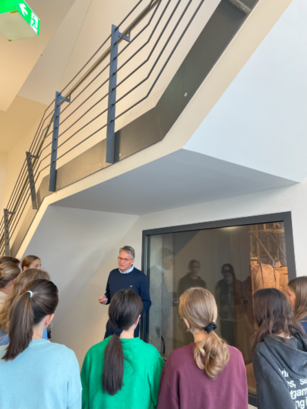 Mitarbeiter spricht mit einer Gruppe von Girls' Day-Teilnehmerinnen in einem modernen Innenraum mit Treppe und Glaswand