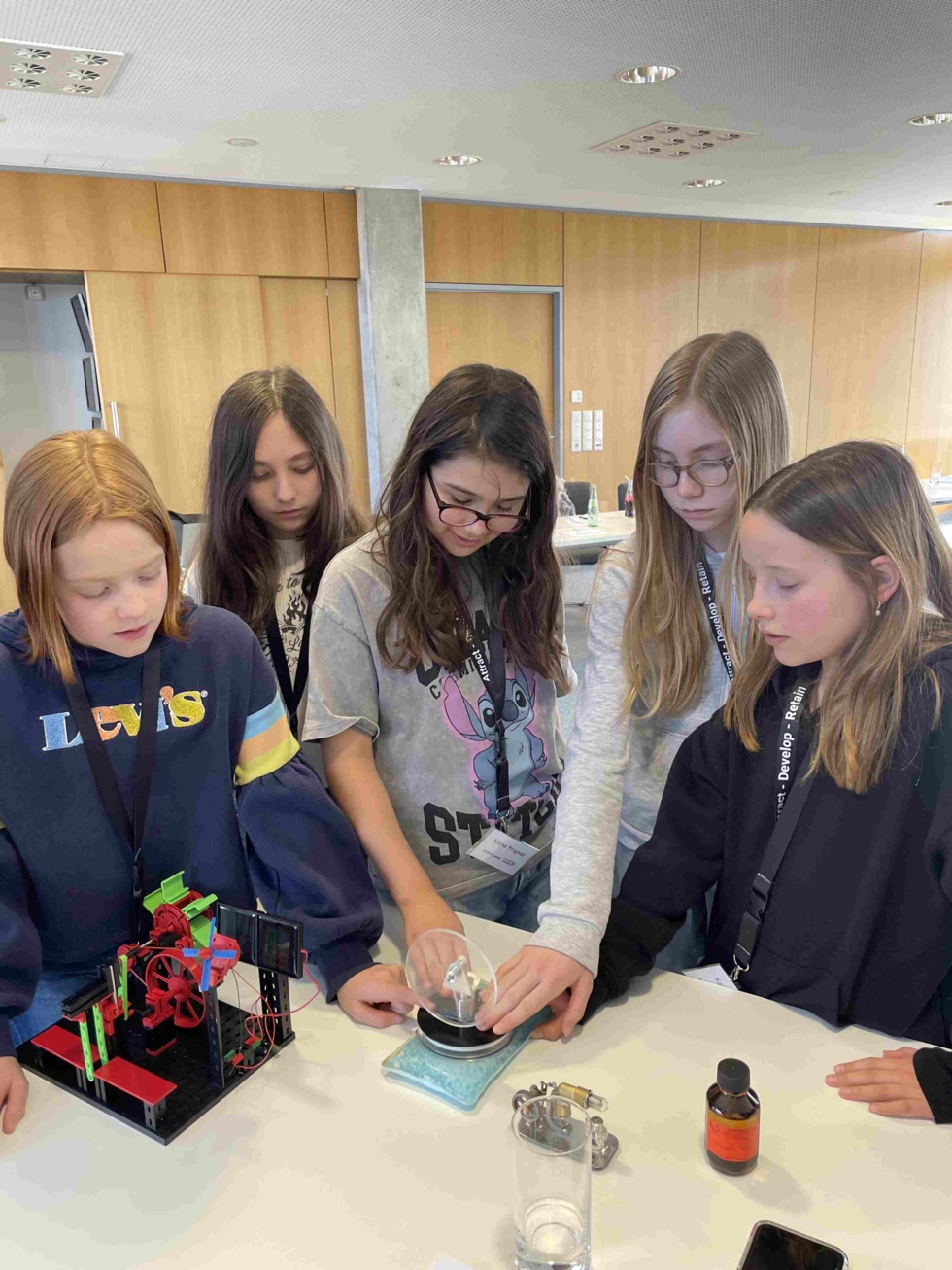 Fünf Girls' Day-Teilnehmerinnen stehen um einen Tisch und experimentieren mit einem Glas und einer Waage, neben ihnen ein kleines technisches Modell und eine Flasche mit Flüssigkeit