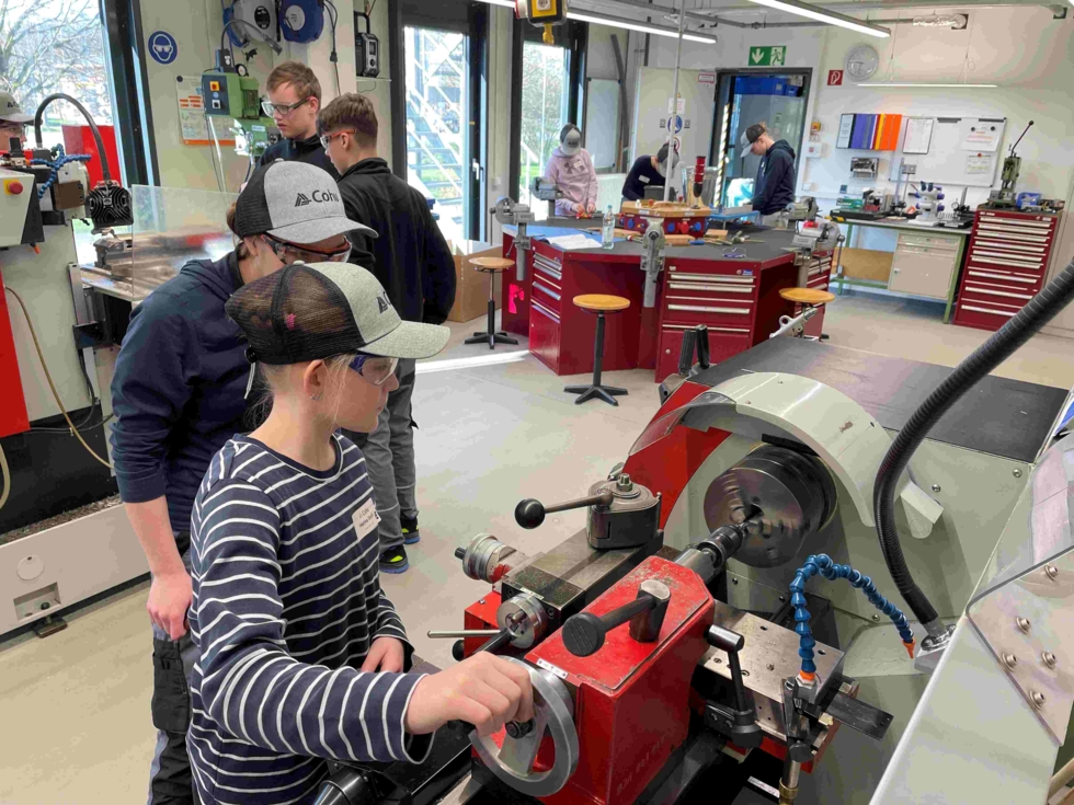 Girls' Day-Teilnehmerinnen in Werkstatt bedienen Drehmaschine, tragen Schutzbrillen und Kappen, andere arbeiten an Werkbänken im Hintergrund