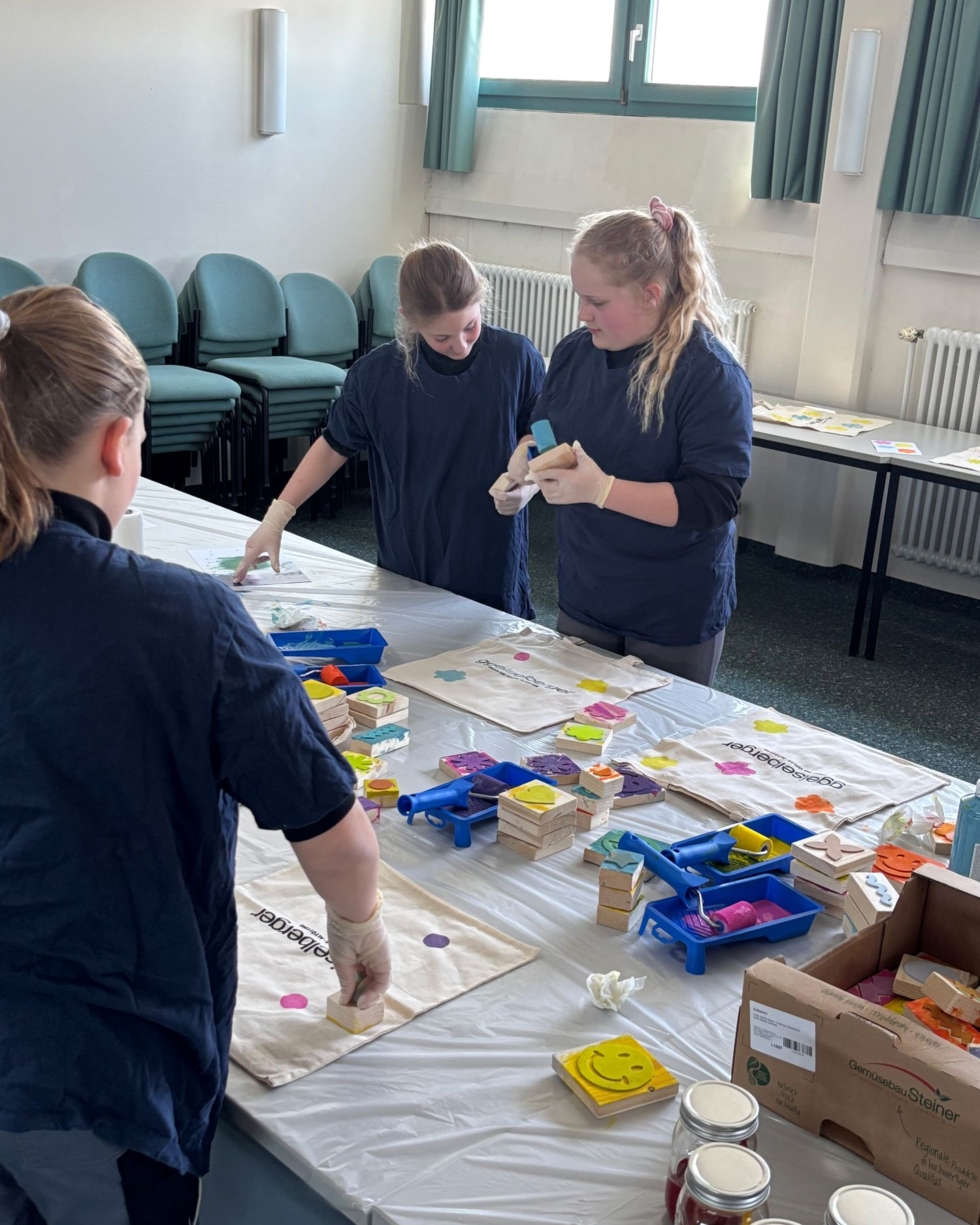 Drei Girls' Day-Teilnehmerinnen arbeiten an einem Tisch mit Bastelmaterialien wie Holzklötzen, Farben und Pinseln in einem hellen Raum mit grünen Vorhängen