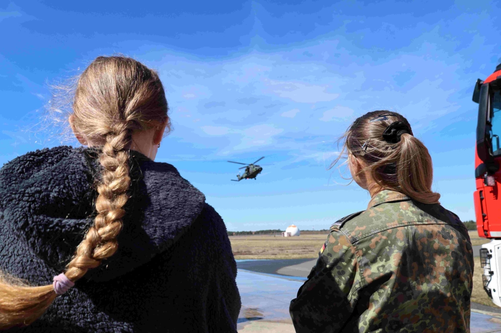 Eine Soldatin und ein Mädchen von hinten fotografiert beobachten einen Hubschrauber.