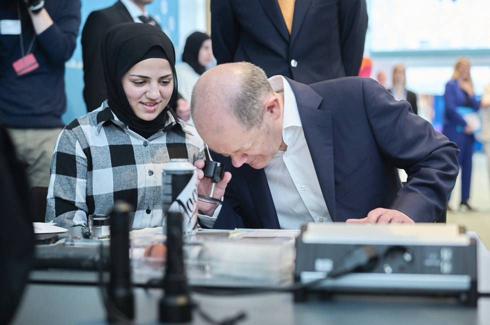 Olaf Scholz guckt durch eine Lupe, eine Schülerin sitzt neben ihm.