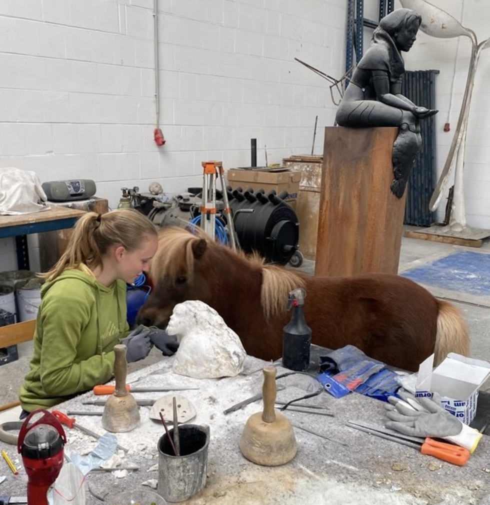 Mädchen bearbeitet einen Stein, ein Pony steht im Hintergrund