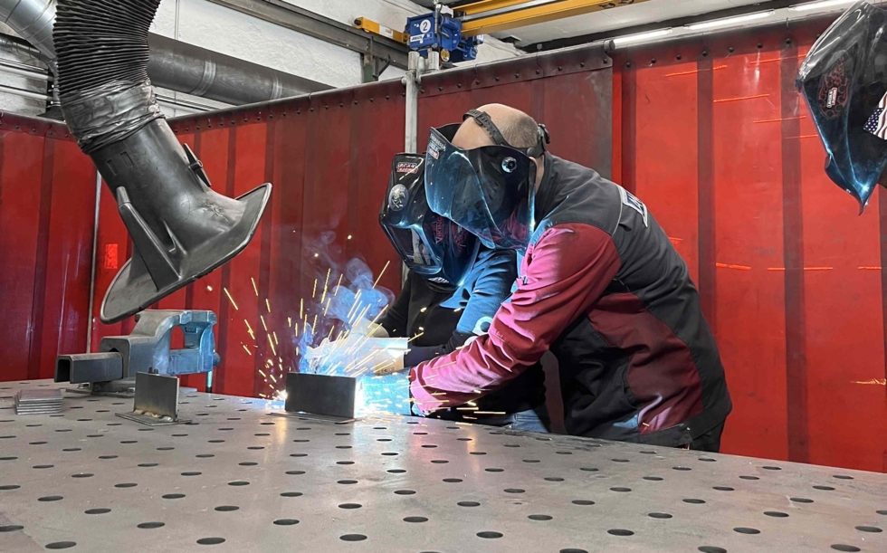 Zwei Personen beim Schweißen in einer Werkstatt, Funken fliegen, Schutzhelme und -kleidung getragen.