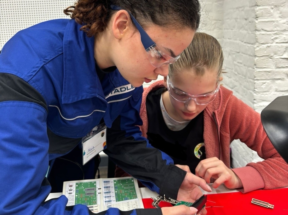 Girls' Day-Teilnehmerin lernt das Löten. Eine Mitarbeiterin zeigt ihr, wie man mit Werkzeugen arbeitet, beide tragen Schutzbrillen.