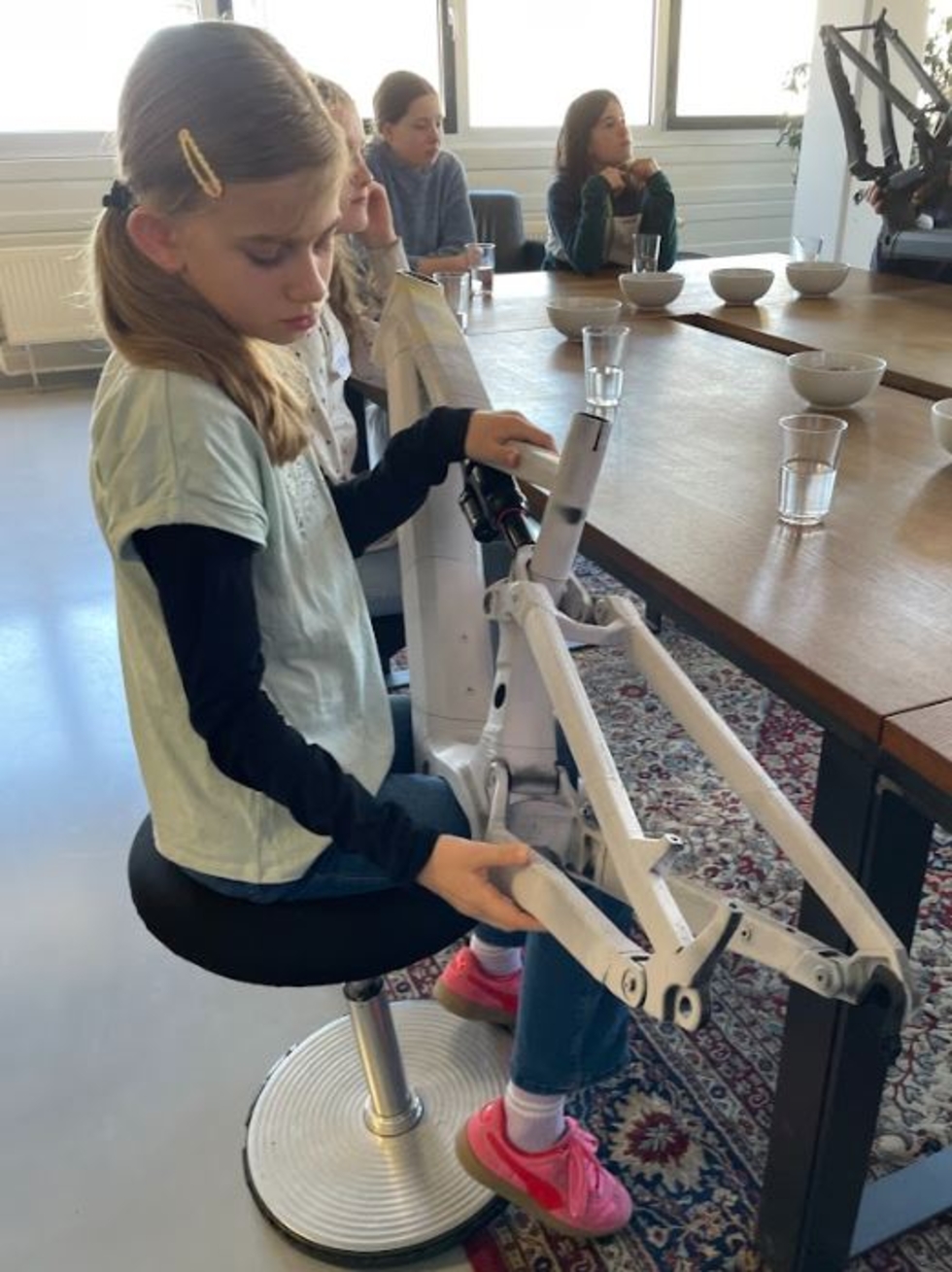 Girls' Day-Teilnehmerin sitzt auf einem Hocker und hält einen weißen Roboterarm, im Hintergrund sitzen weitere Girls' Day-Teilnehmerinnen an einem Tisch mit Gläsern und Schalen
