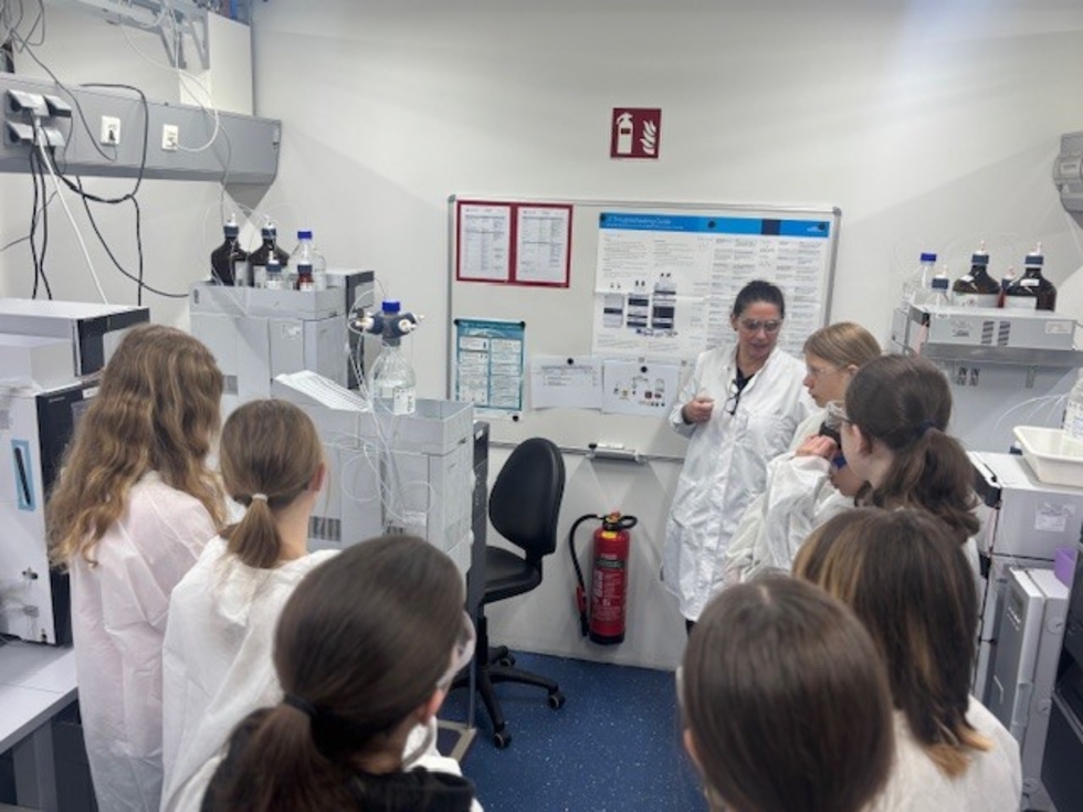 Girls' Day-Teilnehmerinnen und eine Mitarbeiterin in weißen Laborkitteln stehen in einem Laborraum, die Mitarbeiterin erklärt etwas an einem Gerät