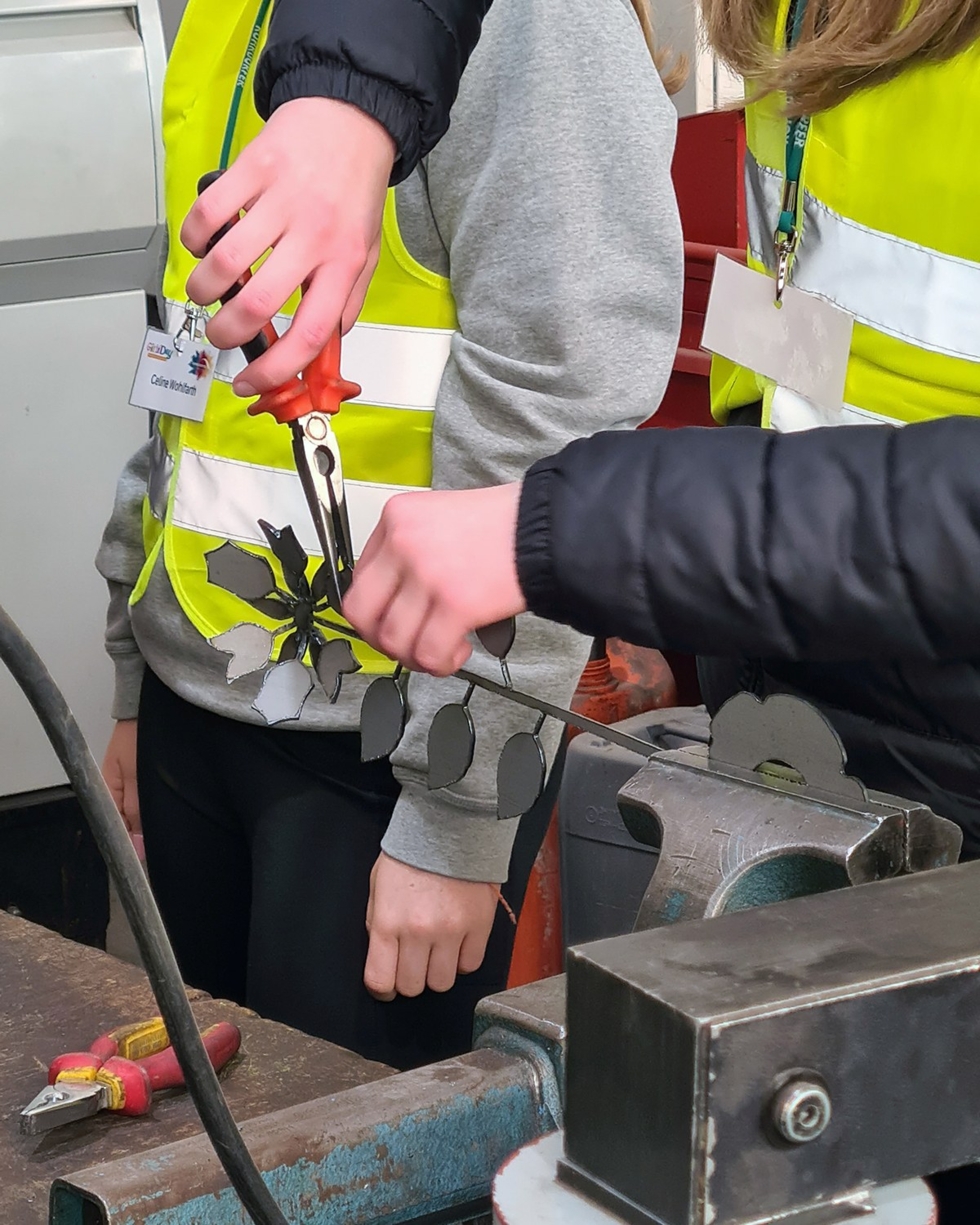Zwei Girls' Day-Teilnehmerinnen in Sicherheitswesten arbeiten gemeinsam an einer Metallbearbeitung, eine hält eine Schere, während die andere ein Werkstück hält.