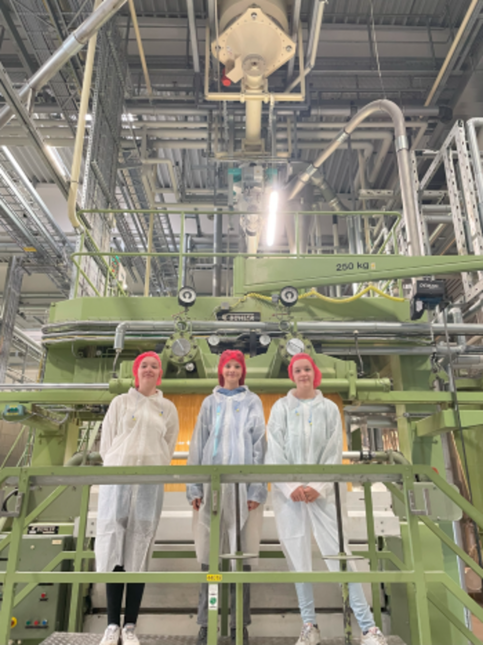 Drei Girls' Day-Teilnehmerinnen stehen in Schutzkleidung und roten Haarnetzen vor einer großen industriellen Maschine in einer Fabrikhalle