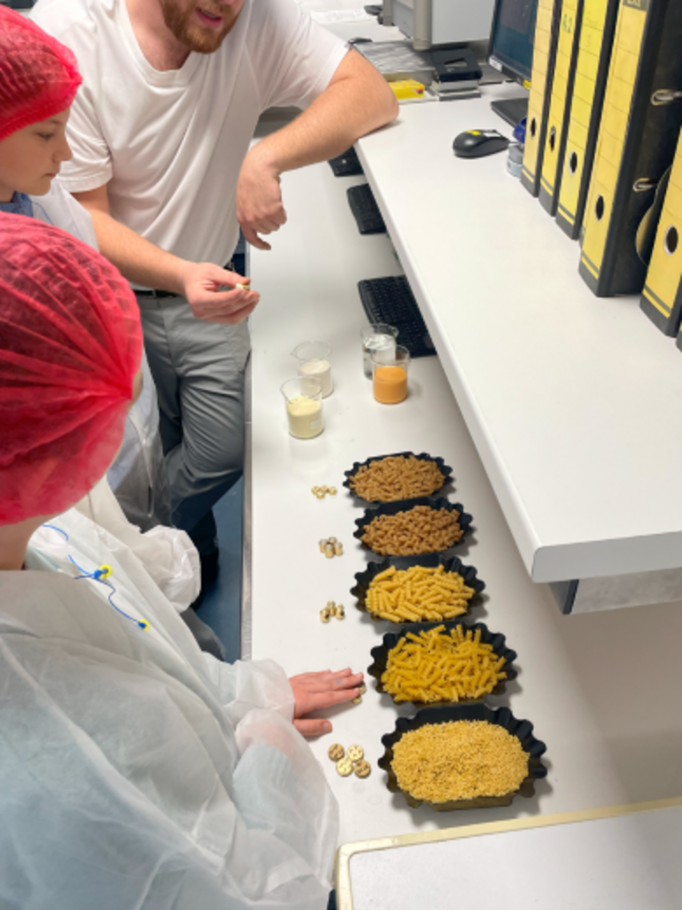 Zwei Girls' Day-Teilnehmerinnen in Schutzkleidung und Haarnetzen betrachten verschiedene Sorten von Nudeln und Gläser mit Flüssigkeiten auf einem Tisch in einem Labor oder einer Produktionsumgebung