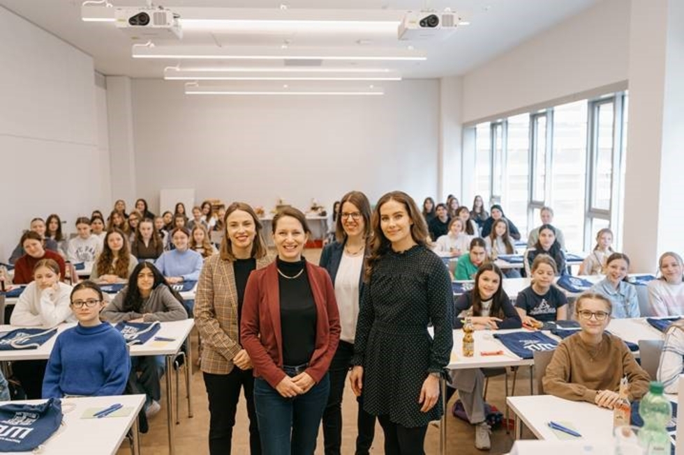 Vier Mitarbeiterinnen stehen vorne in einem Raum voller Girls' Day-Teilnehmerinnen, die an Tischen sitzen