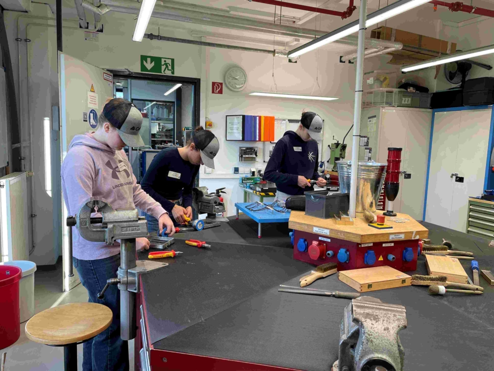 Drei Girls' Day-Teilnehmerinnen arbeiten mit Schutzbrillen an Werkbänken in einer Werkstatt mit Werkzeugen und Maschinen