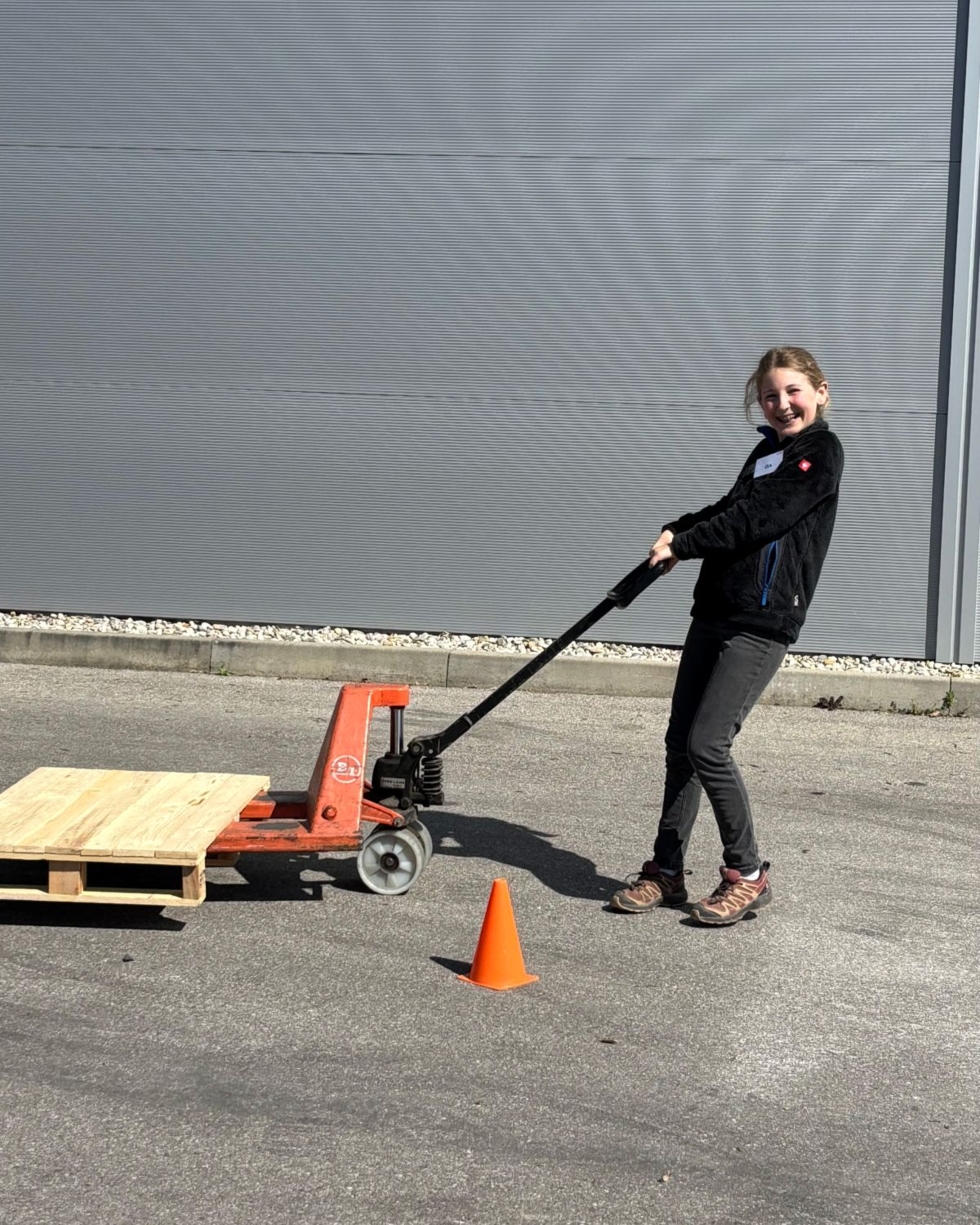 Eine Girls' Day-Teilnehmerin zieht Hubwagen mit leerer Holzpalette auf Asphalt vor grauer Wand, orangefarbener Verkehrskegel daneben