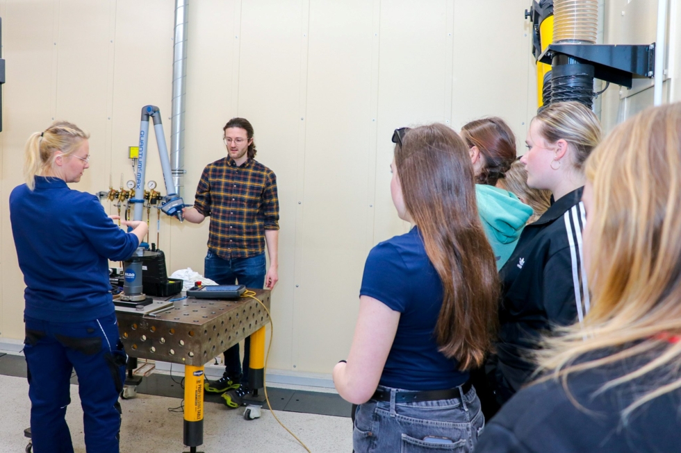 Mitarbeitende erklären Girls' Day-Teilnehmerinnen in einem Werkraum die Funktion eines technischen Geräts auf einem Arbeitstisch