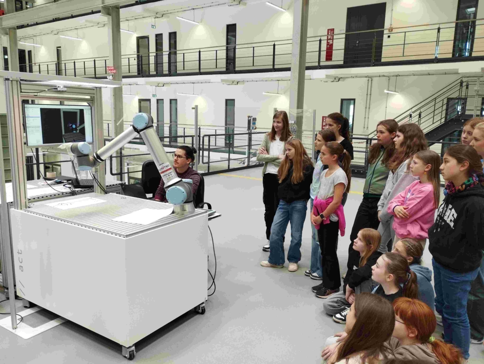 Gruppe von Girls' Day-Teilnehmerinnen beobachtet einen Mitarbeiter, der einen Roboterarm in einem modernen Labor oder Technikraum bedient