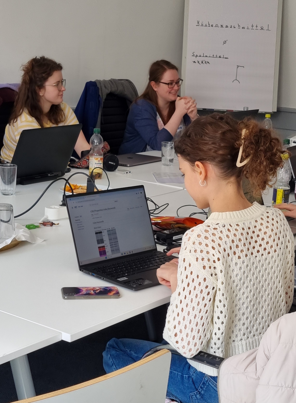 Drei Girls' Day-Teilnehmerinnen sitzen an einem Tisch mit Laptops und Getränken, im Hintergrund ein Flipchart mit handgeschriebenem Text