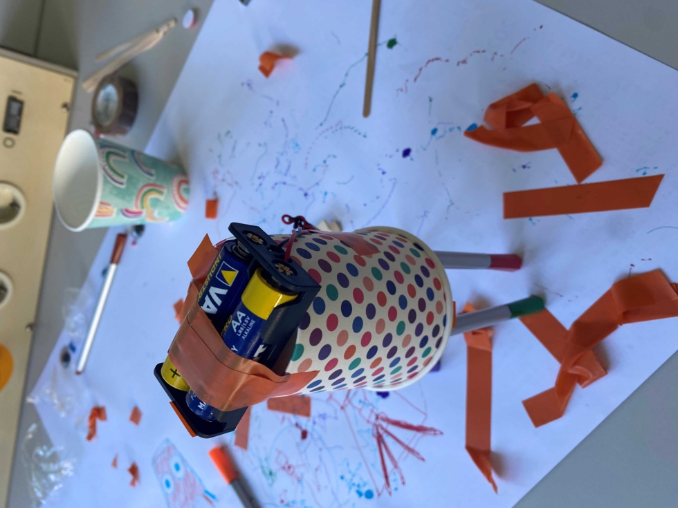 Bastelprojekt mit Pappbecher, Batterien und orangem Klebeband auf weißem Papier mit Kritzelzeichnungen