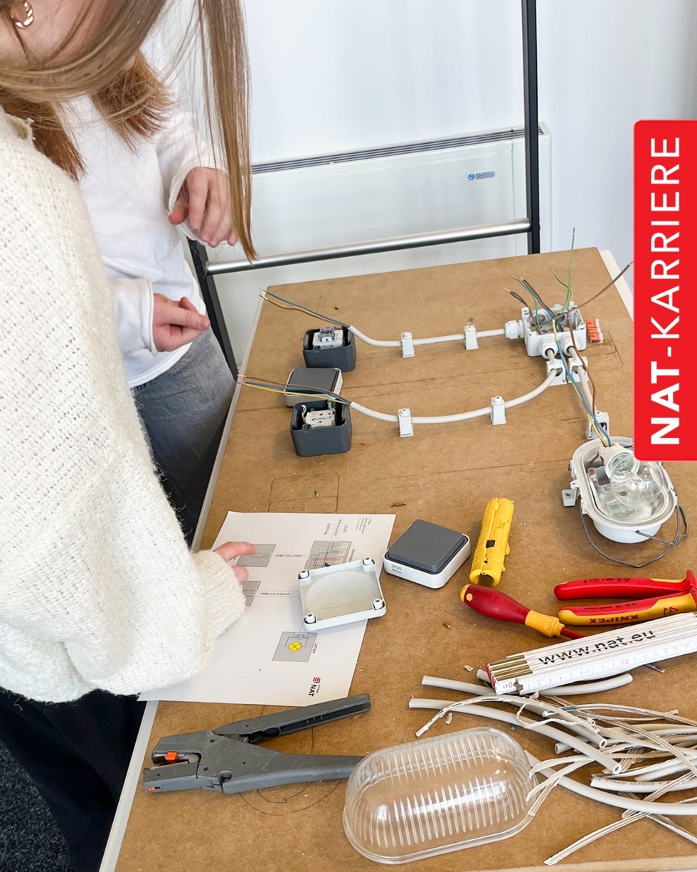Zwei Girls' Day-Teilnehmerinnen arbeiten an einem Tisch mit elektrischen Kabeln, Werkzeugen und Schaltplänen