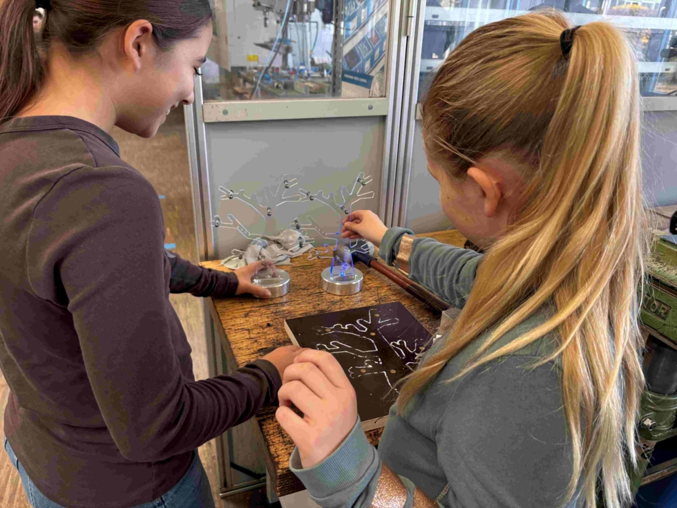 Zwei Girls' Day-Teilnehmerinnen experimentieren mit einem kleinen elektrischen Gerät, das blaue Funken erzeugt, auf einem Labortisch