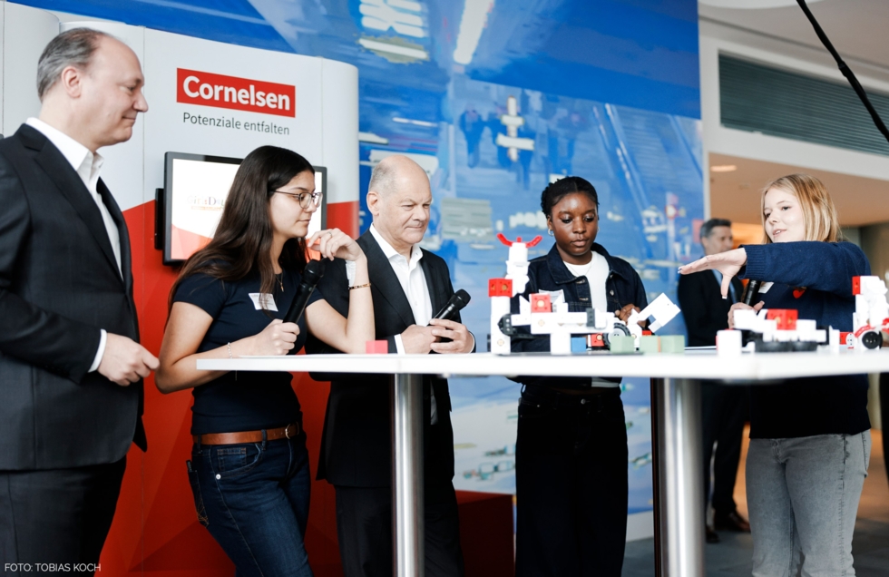Drei Schülerinnen präsentieren Bundeskanzler Olaf Scholz am Stand von Cornelsen beim Girls'Day-Auftakt 2025 ein Technikprojekt mit Lernrobotern. Eine Schülerin erklärt gerade aktiv mit ausgestreckter Hand, während die anderen aufmerksam zuschauen.