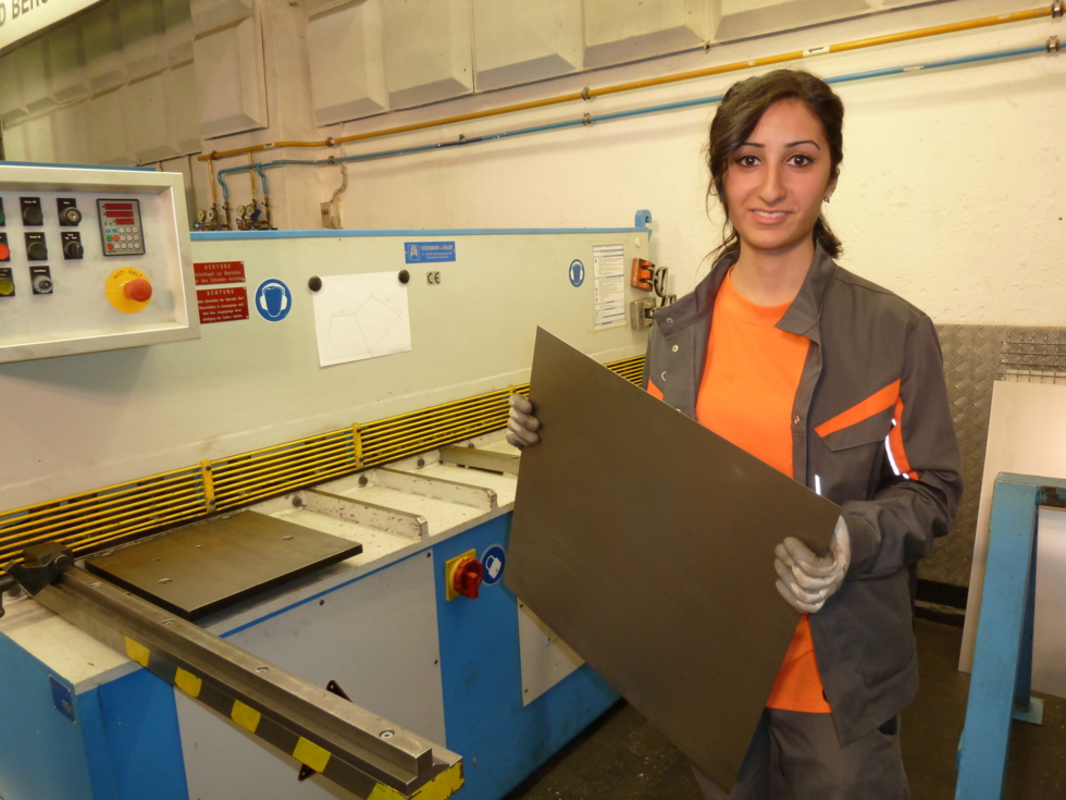 Junge Frau hält vor einer Werkbank eine Metallplatte in der Hand