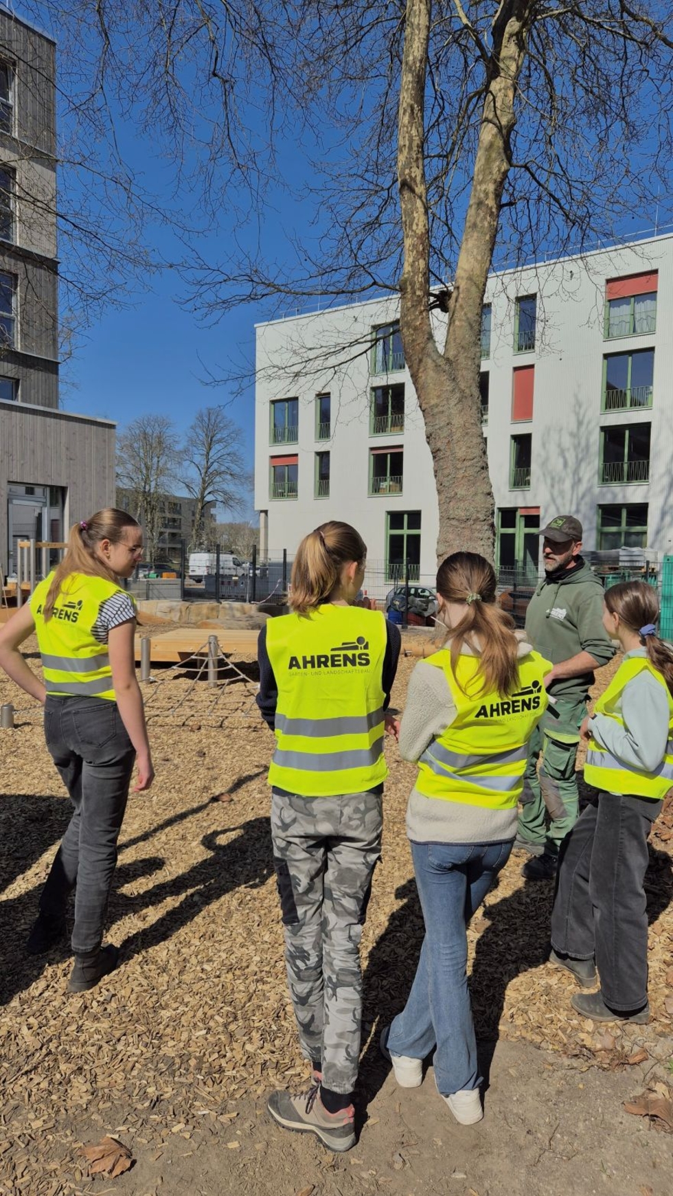 Vier Girls' Day-Teilnehmerinnen und ein Mitarbeiter mit gelben Warnwesten stehen im Freien vor einem Baum und Gebäuden, sie scheinen eine Besprechung zu haben