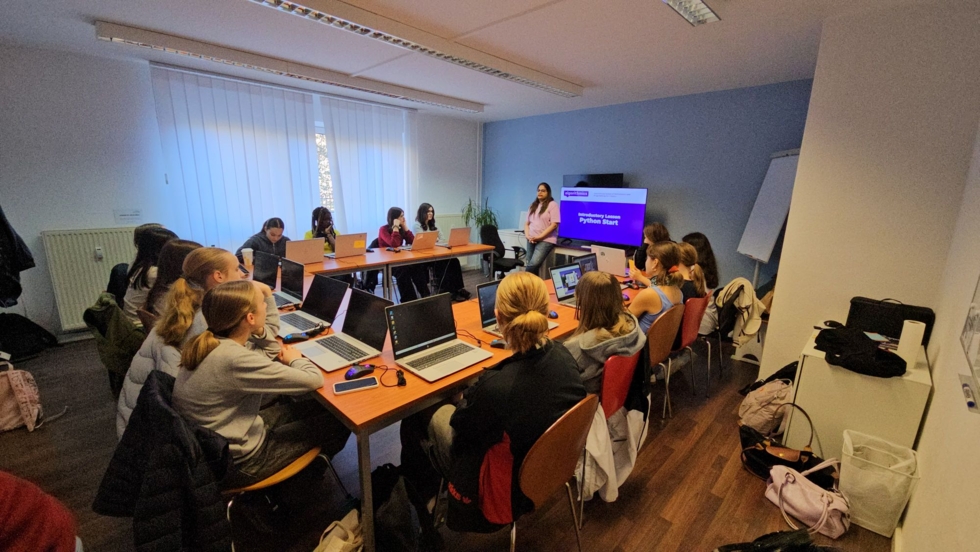 Girls' Day-Teilnehmerinnen sitzen in einem Besprechungsraum an einem langen Tisch mit Laptops, während eine Mitarbeiterin vor einem Bildschirm mit Präsentation steht
