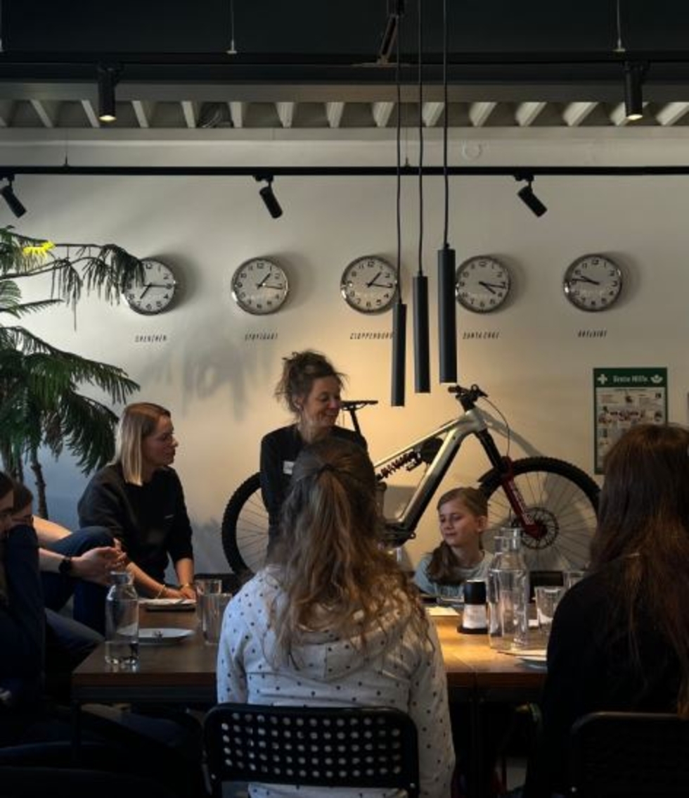 Mitarbeiterinnen und Girls' Day-Teilnehmerinnen sitzen an einem Tisch in einem Raum mit fünf Wanduhren, die verschiedene Städte zeigen, und einem Fahrrad an der Wand